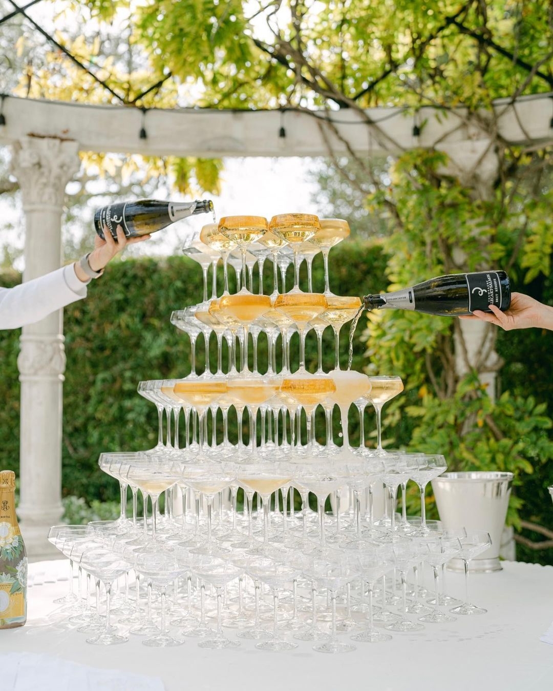 Two people are pouring champagne from a magnum bottle into a pyramid of champagne flutes. The champagne tower is on a table decorated with gold flakes and ivy. There is a black tablecloth in the background.