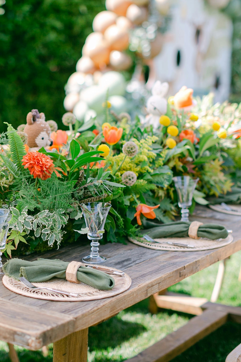 A long picnic table with a centerpiece of colorful flowers running down the middle. The table is set with plates, napkins, and glasses. There are trees and a grassy area in the background.